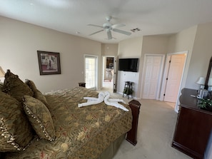 A bedroom with a large bed, a ceiling fan, a TV mounted on the wall, and a door leading to another room.