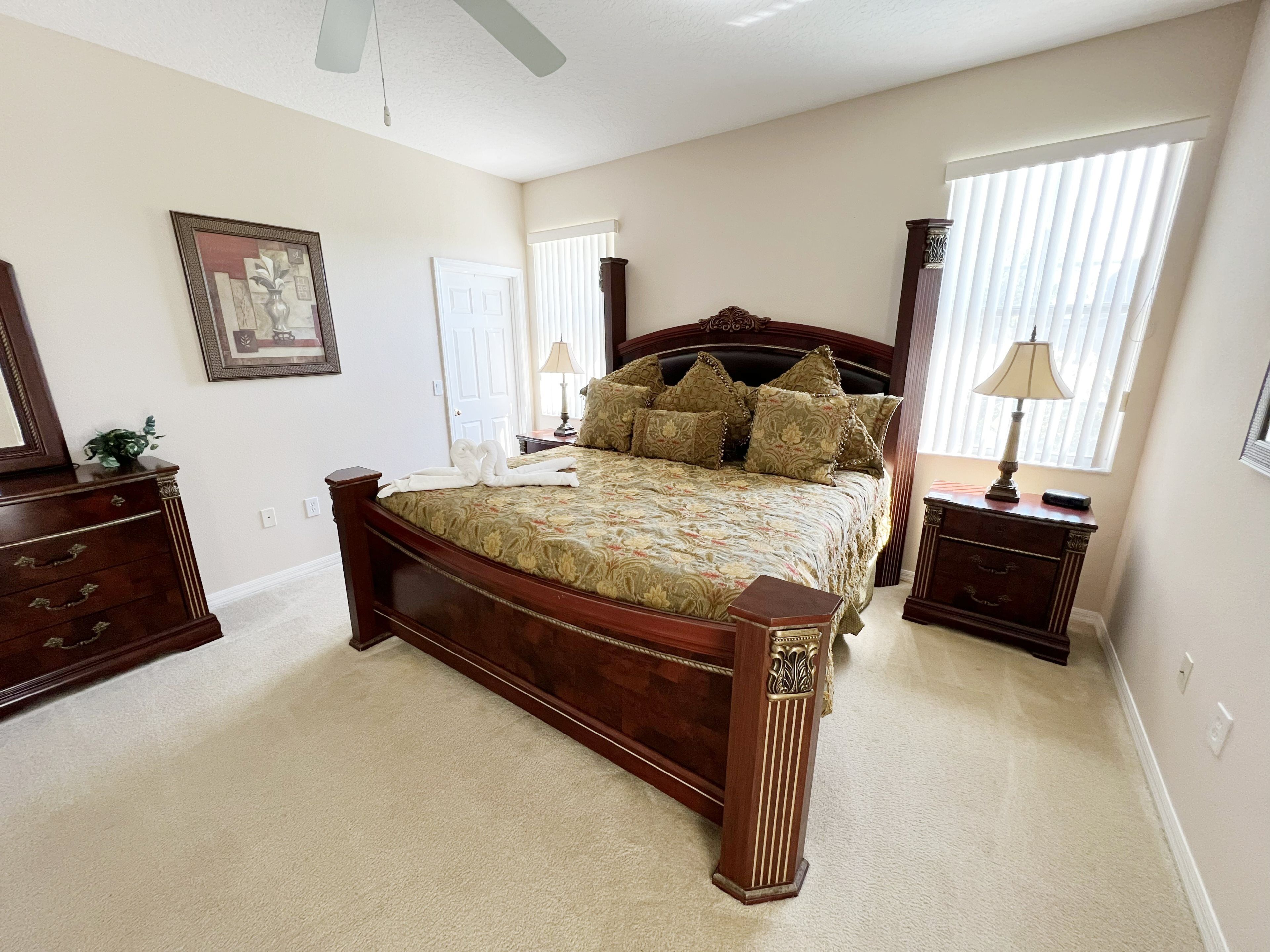A bedroom with a large bed, a dresser, a nightstand, a lamp, a window with blinds, and a ceiling fan.