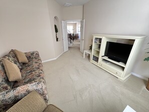 A living room with a floral sofa, a television on a stand, and a hallway leading to a dining area. - Gated Community. Luxury Home 5 Bedrooms/Pool/Spa/Games Room & Den. (Haines City)
