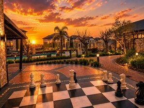 Un jeu d’échecs installé dans une cour, avec une vue sur le coucher de soleil, entouré de bâtiments et d’une piscine.