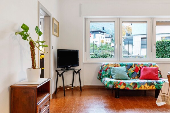 A living room with a sofa, a television, a potted plant, and a window with a view of buildings.