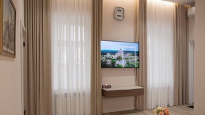 Hotelkamer met een flatscreen televisie aan de muur, een houten console tafel, beige gordijnen, een klein rond tafeltje met een bloemenvaas en een klok.