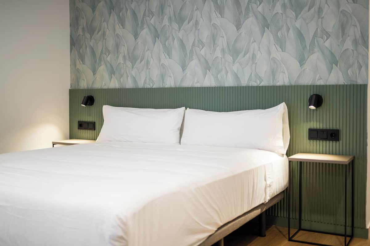 A neatly made bed with white linens in a room with a green-striped headboard and a floral-patterned wallpaper.