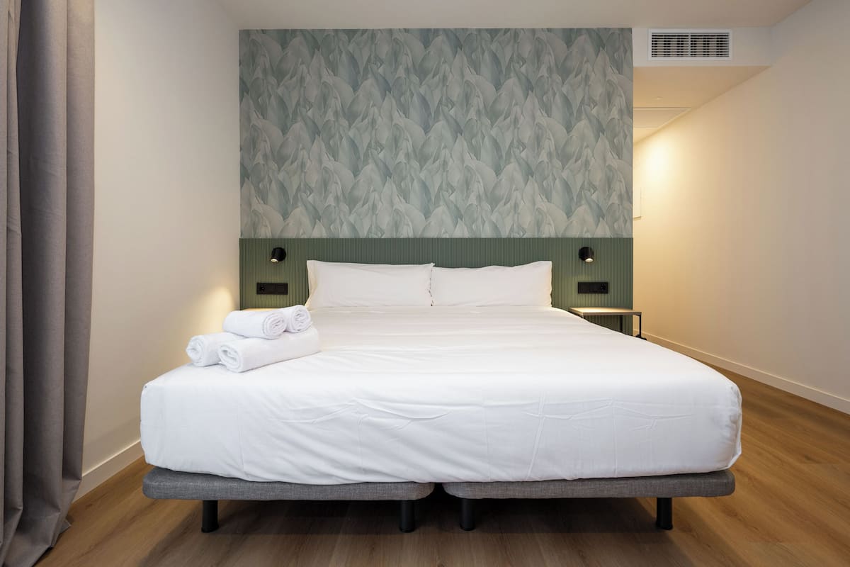 A neatly made bed with white linens, a headboard with a patterned wallpaper, and a bedside table with a lamp.