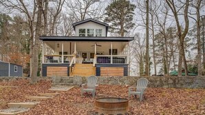 Een modern huis met twee verdiepingen, voorzien van een stenen keerwand, een vuurkorf en twee Adirondack-stoelen in een bebost gebied.