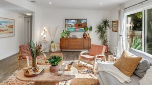 Una sala de estar moderna con un sofá, dos sillones, una mesa de centro y una mesa de comedor. Hay un mueble de madera con adornos y un cuadro en la pared.