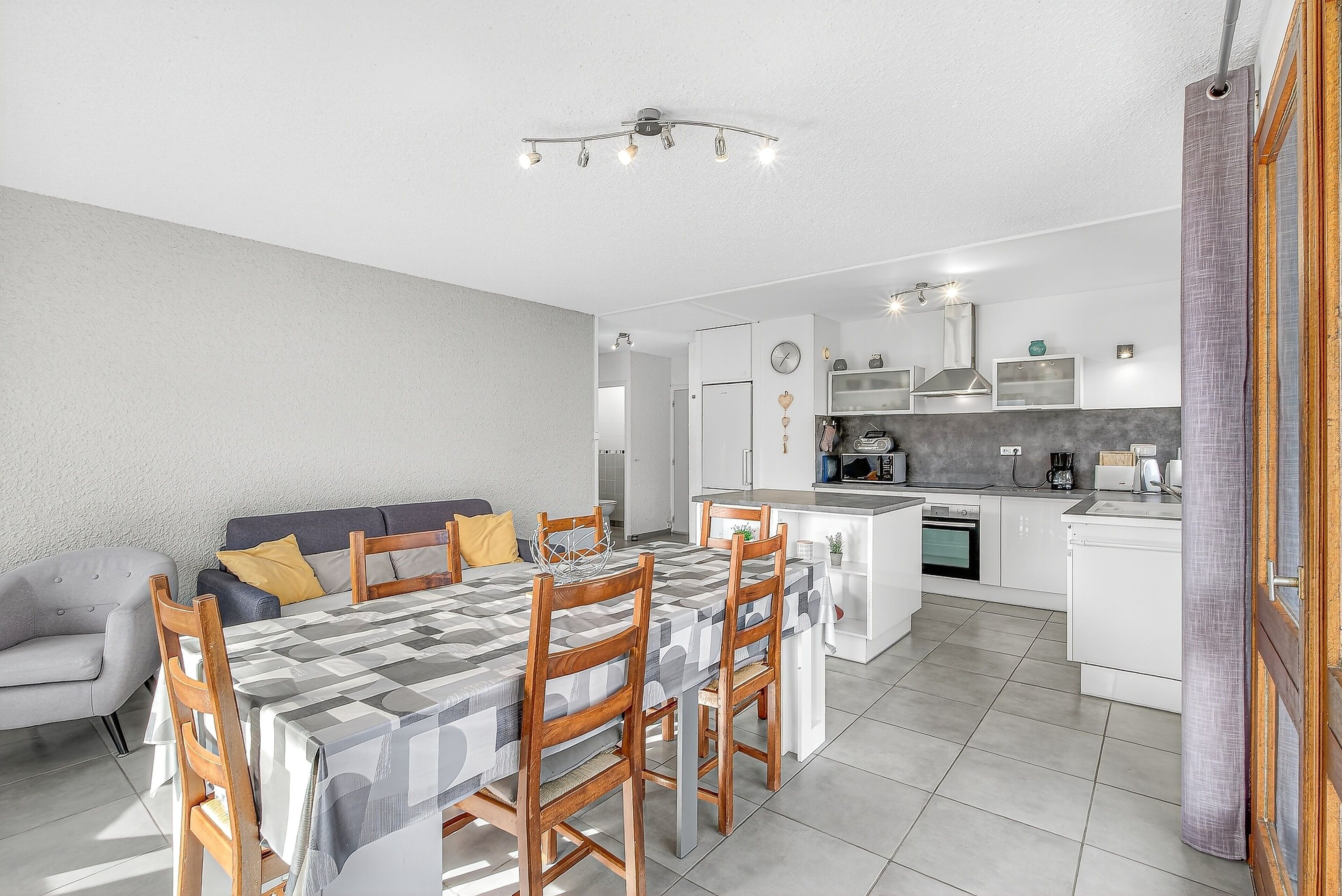 Une cuisine moderne avec des meubles blancs, un coin repas avec une table et des chaises, et un salon avec un canapé et des coussins.