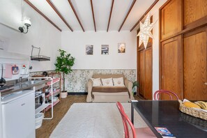 Een kleine keuken met een spoelbak, aanrecht en opslagplanken. Een bank en een eettafel met stoelen staan in de woonkamer.