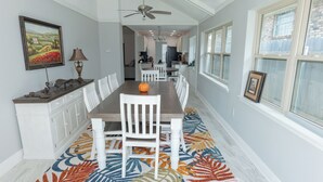 A dining room with a long table, white chairs, a ceiling fan, and a patterned rug.