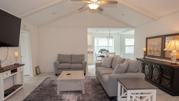A spacious living room with a high ceiling, a ceiling fan, a television mounted on the wall, and a dining area visible through an open doorway.