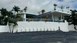 A modern beachfront property with a white exterior, a covered patio, and palm trees.