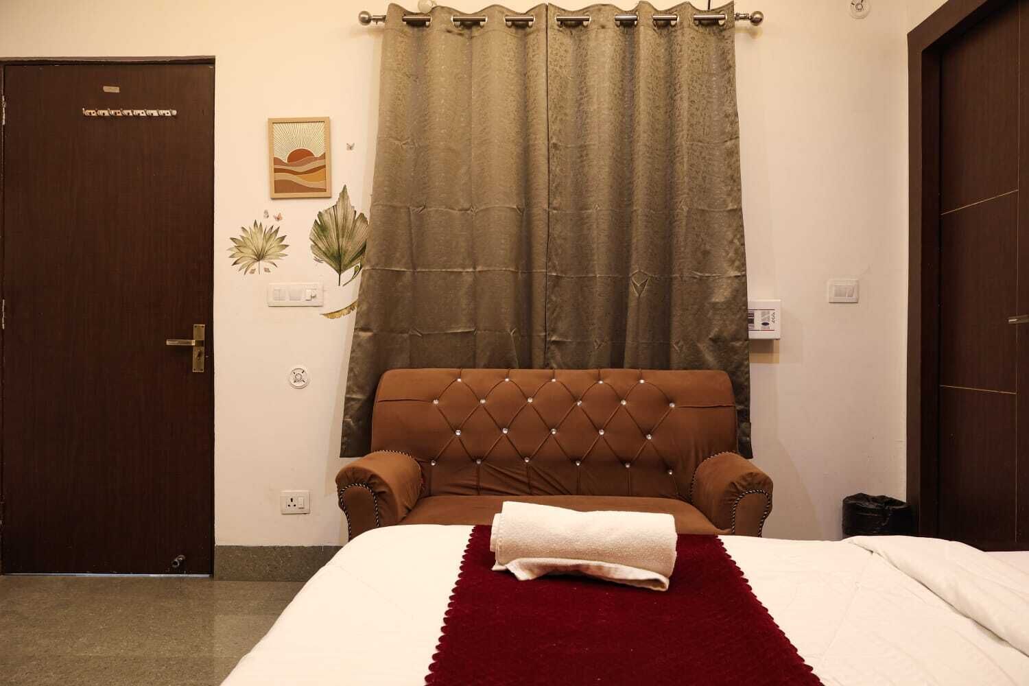 Une chambre à coucher avec un lit, une tête de lit marron recouverte de tissu, un plaid rouge, une porte marron et un rideau.