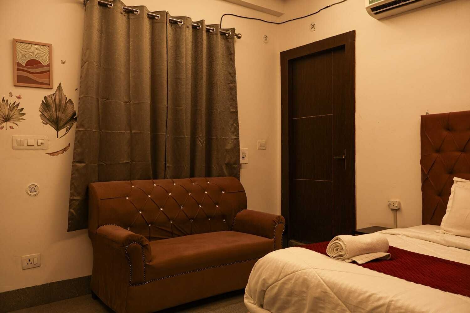 A bedroom with a brown sofa, a bed with white bedding, a dark wooden door, and a wall-mounted air conditioner.