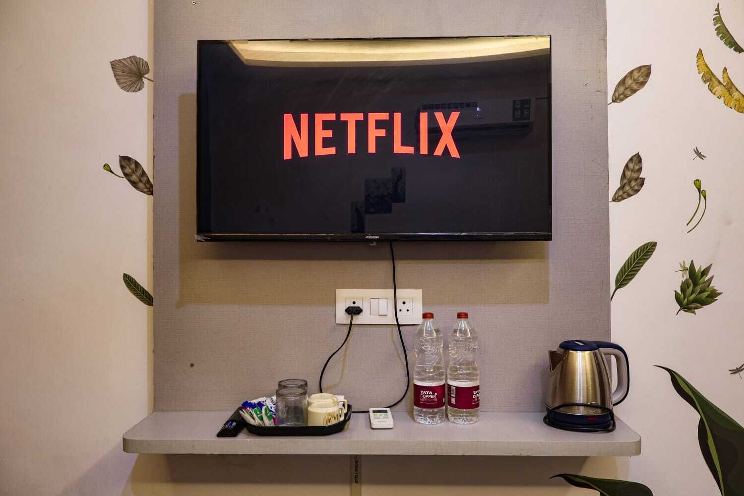 Ein an der Wand befestigter Fernseher mit Netflix-Logo, eine Steckdose, eine Ablage mit einer Tasse, zwei Wasserflaschen und eine Kaffeemaschine.