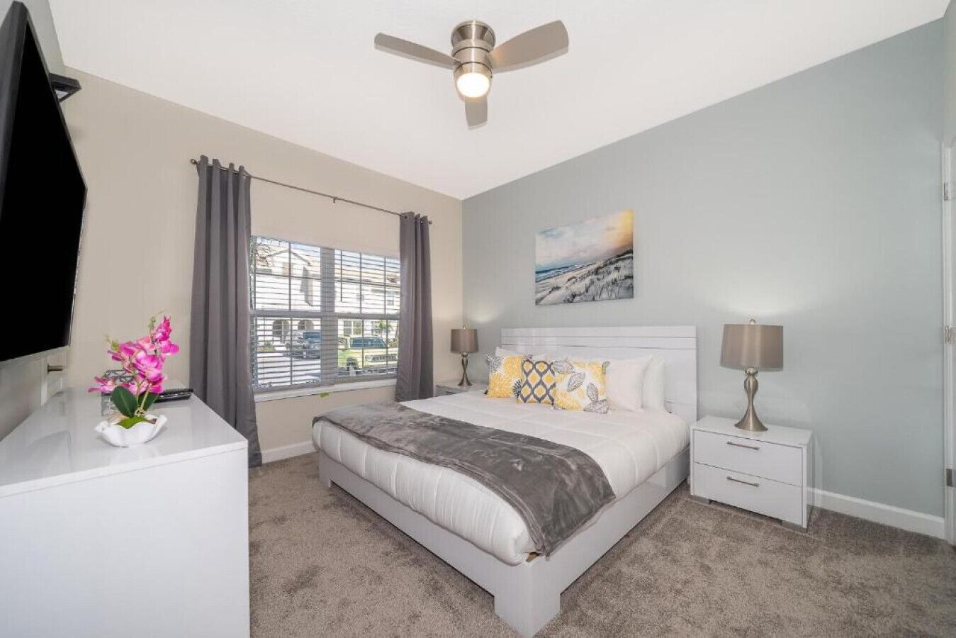 A bedroom with a bed, bedside tables, a ceiling fan, a window with curtains, and a painting on the wall.