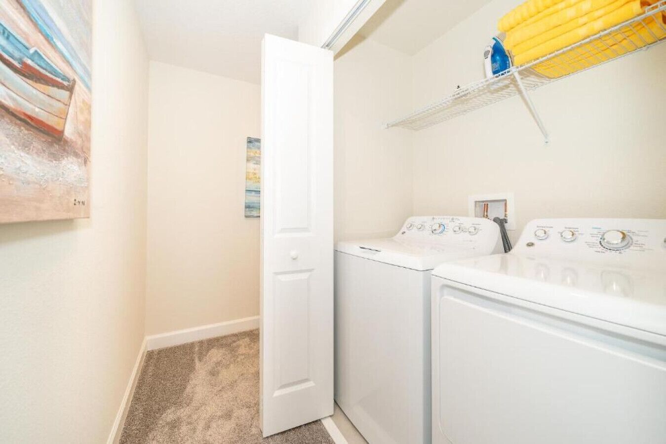 A laundry room with a white door, a washing machine, and a dryer. There is a shelf with yellow towels and a painting on the wall.