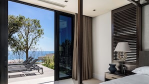 A modern hotel room with a large sliding glass door leading to a balcony, a bed with a headboard, a bedside table with a lamp, and a view of the sea. - Halcyon Villa (Cape Town)