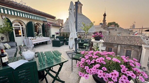 Een terras op het dak met buitenmeubilair, een tafel met glazen en een bloemstuk.