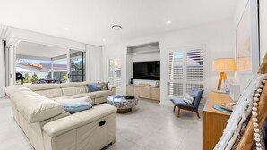 A modern living room with a beige sofa, a coffee table, and a TV. There is a sliding glass door leading to an outdoor area with a barbecue.
