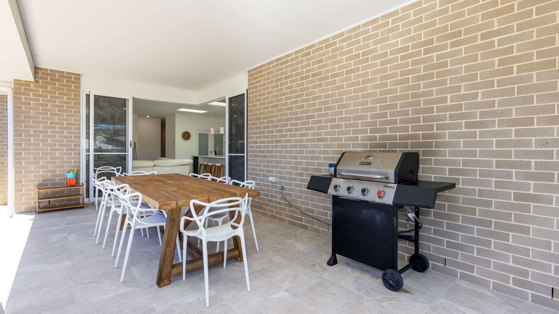 An outdoor patio with a wooden table and white chairs, a barbecue grill, and a brick wall.