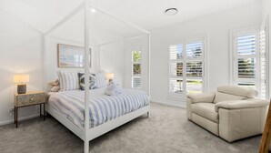 A bedroom with a four-poster bed, a nightstand, a chair, and large windows with blinds.