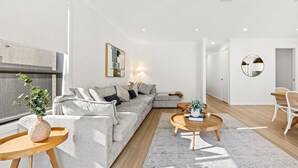 A modern living room with a grey sectional sofa, a round wooden coffee table, and a dining area with wicker chairs. - Callala Chill Jervis Bay - Short Walk to Creek (Callala Bay)