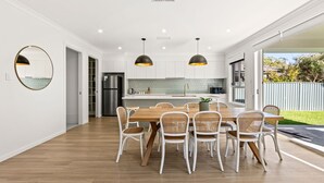 A modern kitchen with a dining area, featuring a wooden table and wicker chairs, a built-in oven, and pendant lighting. - Callala Chill Jervis Bay - Short Walk to Creek (Callala Bay)