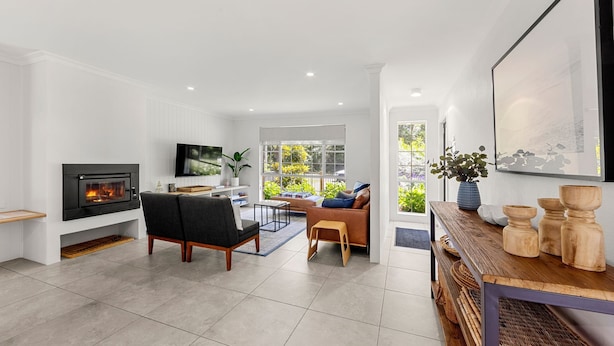 A modern living room with a fireplace, a sofa, a coffee table, and a TV. There is a window with a view of greenery outside. - Callala Bay n Beach House - Pet Friendly with Fireplace (Callala Bay)