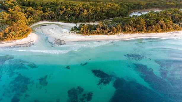 Oversigtsbillede af et kystområde med krystalklart turkis vand, sandstrande og frodig, grøn vegetation.