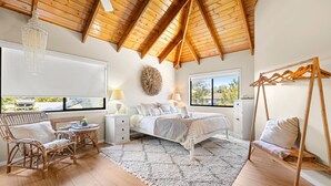 Un dormitorio con techo de madera, una cama con ropa blanca, una silla de mimbre, una mesita con una lámpara y vistas al exterior a través de una ventana.