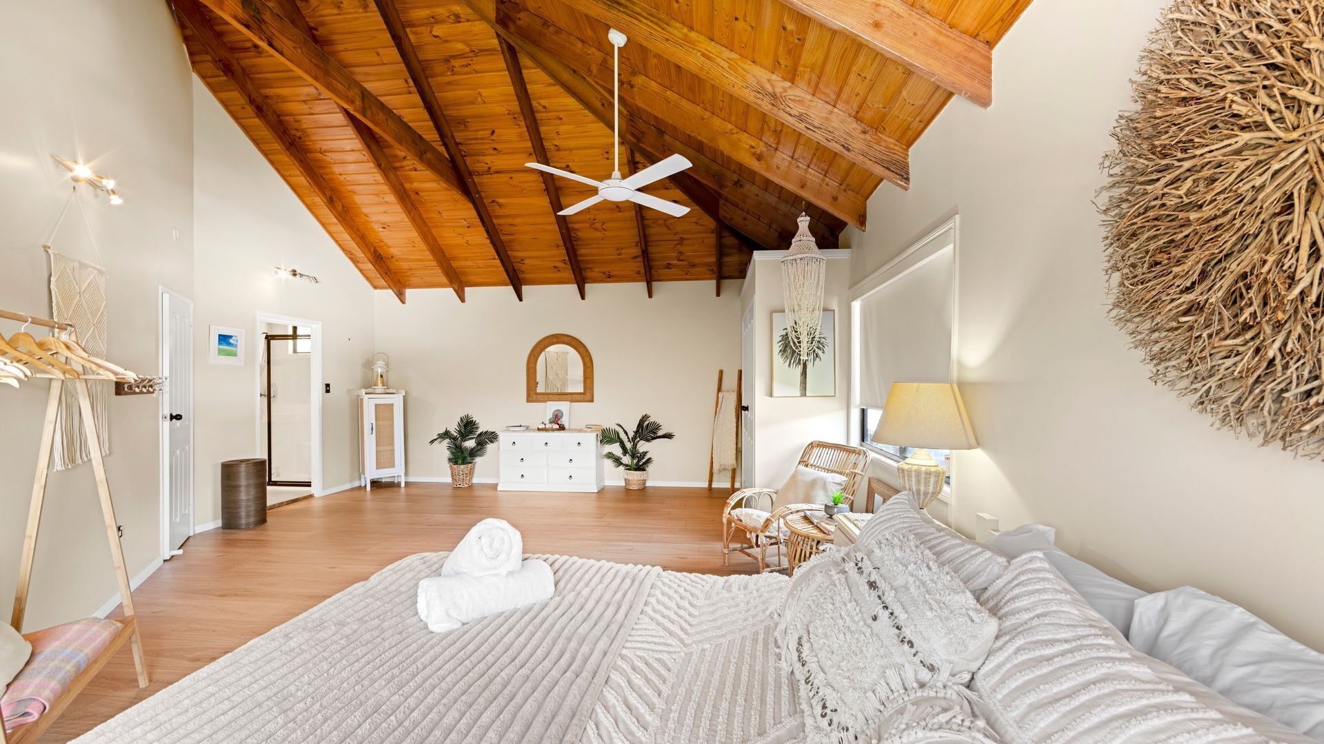 Amplia habitación con techo alto de madera, ventilador de techo, cama con colcha blanca, tapiz grande tejido en la pared y cómoda blanca con plantas.
