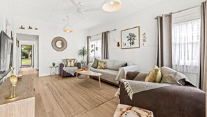 A modern living room with a sofa, a coffee table, and a TV. There is a dining table with chairs, a plant, and a decorative mirror. The room has a ceiling fan and a window with curtains. - Osprey - Pet Friendly Waterfront (Callala Bay)
