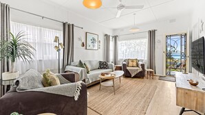 A spacious living room with a sofa, armchair, coffee table, and a TV. There is a large window with curtains, a ceiling fan, and a view of the outdoors. - Osprey - Pet Friendly Waterfront (Callala Bay)