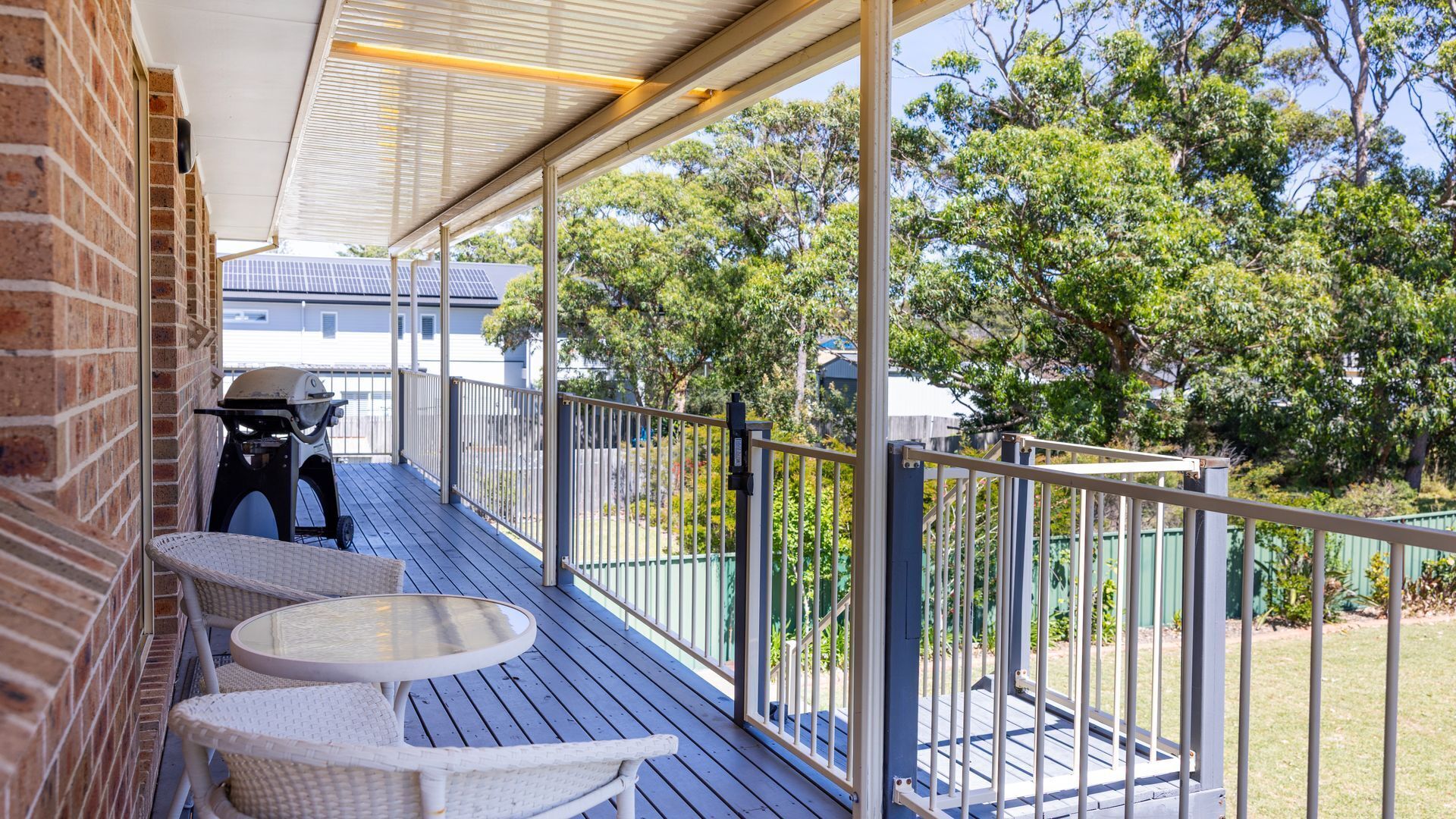 Un patio exterior techado con una parrilla, mobiliario de mimbre y vistas a una zona residencial con árboles y casas.