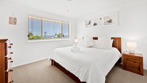 A bedroom with a large bed, a nightstand with a lamp, a dresser, and a window with blinds. - Sandy Adventures - Pet Friendly - 1 Min to Beach (Callala Beach)