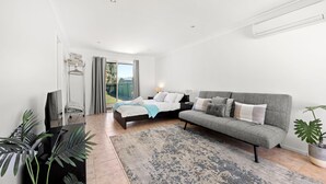 A modern bedroom with a bed, a grey sofa, a TV, and a large window. - Sandy Adventures - Pet Friendly - 1 Min to Beach (Callala Beach)