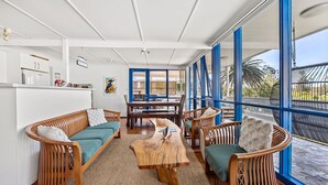 Amplia sala de estar con mesa de centro de madera, sofás de mimbre y una mesa de comedor con sillas. Ventanas grandes ofrecen vistas al exterior.
