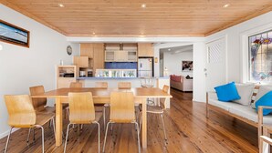 Een moderne keuken met houten kastjes en een centraal eiland, een eetgedeelte met een houten tafel en stoelen, en een gezellige zithoek met een bank en kussens.