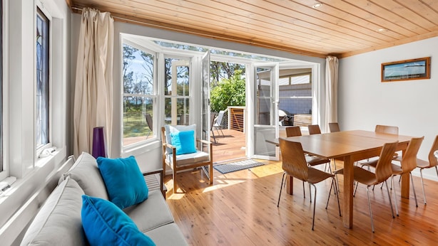 Een moderne woonkamer met een houten vloer, een bank met blauwe kussens, een eettafel met stoelen en grote ramen met uitzicht op het landschap.