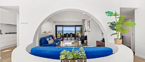 A modern living room with a blue sofa, a coffee table with plants, and a view of the sea through an arched doorway.