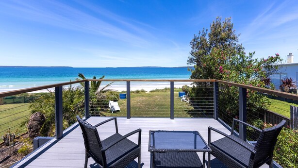 Terraza con dos sillas de mimbre y una mesa de vidrio con vista a una playa, y una vista despejada del océano y colinas distantes.