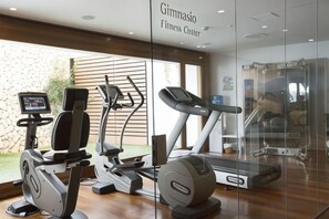 A fitness center with cardio machines, a digital display, and a glass wall. - Hotel Hospes Maricel & Spa (Ca's Catala)