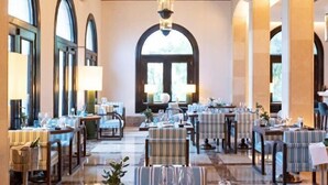 A dining area with tables and chairs, large windows, and a chandelier. - Hotel Hospes Maricel & Spa (Ca's Catala)