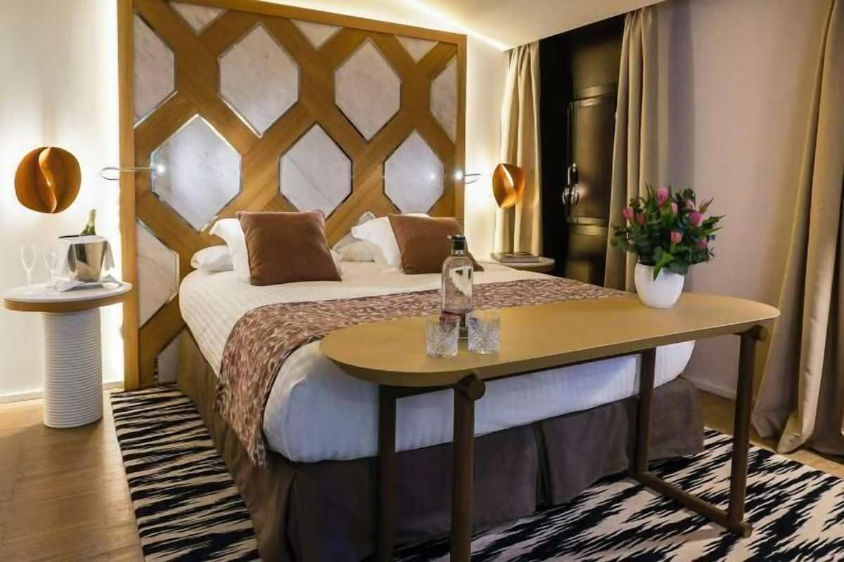 Hotellihuone, jossa on sänky, yöpöytä, seinälle asennettu televisio, ikkuna verhoilla ja ruukussa oleva kasvi.