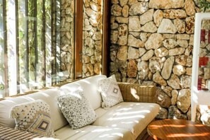 A room with a stone wall, a white sofa with patterned cushions, and a wicker chair. - Hotel Hospes Maricel & Spa (Ca's Catala)