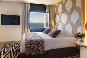 A hotel room with a bed, a view of the sea, a wall-mounted TV, and a patterned rug. - Hotel Hospes Maricel & Spa (Ca's Catala)