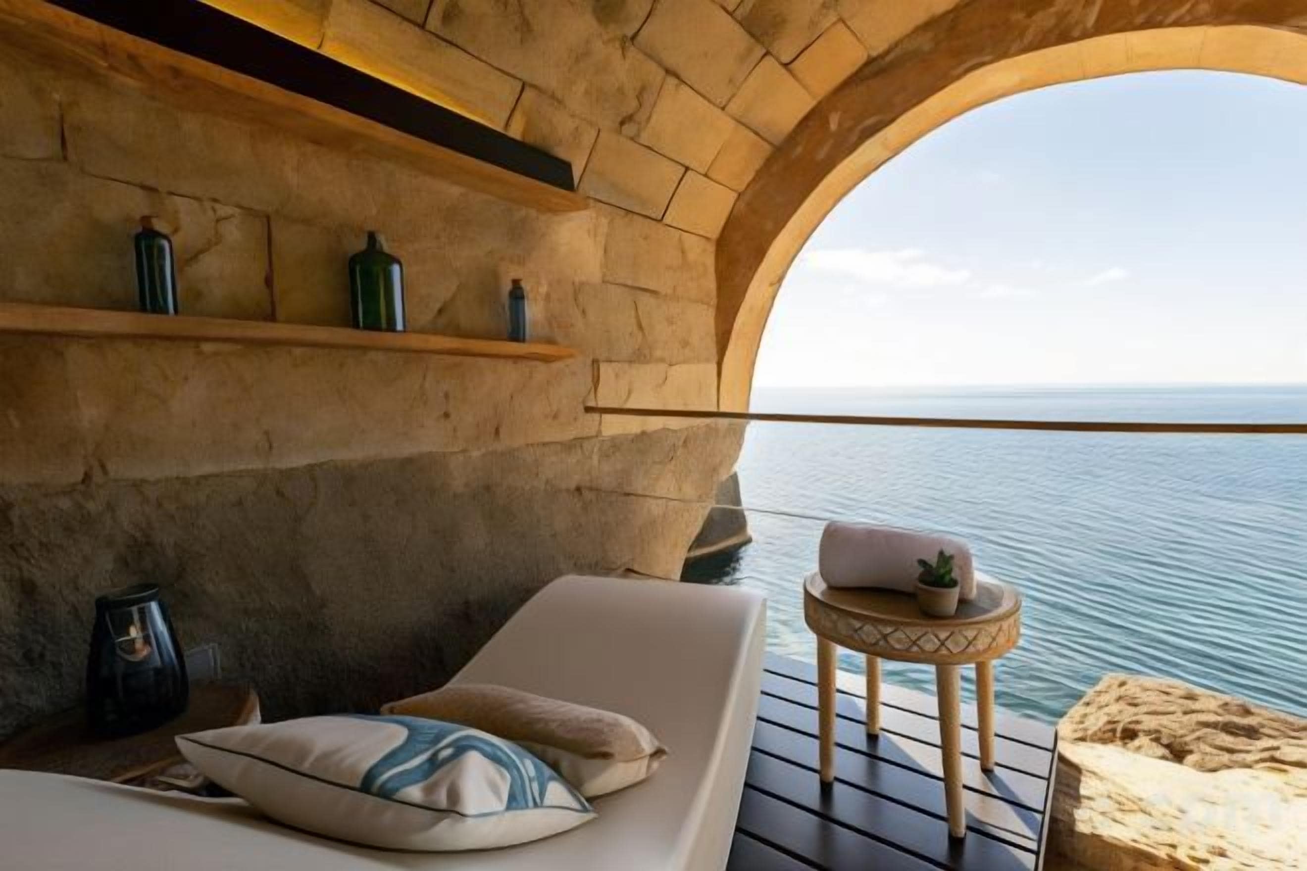 Una habitación con pared de piedra, una cama con almohadas, una pequeña mesa con una planta y vista al mar.