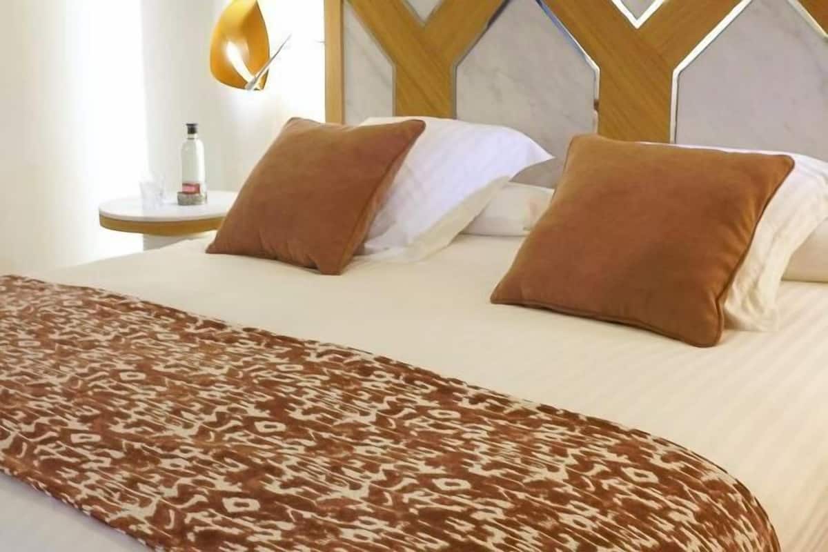 Hotellihuone, jossa on sänky, ruskea kuviollinen päiväpeite, valkoiset tyynyt ja puinen pääty.