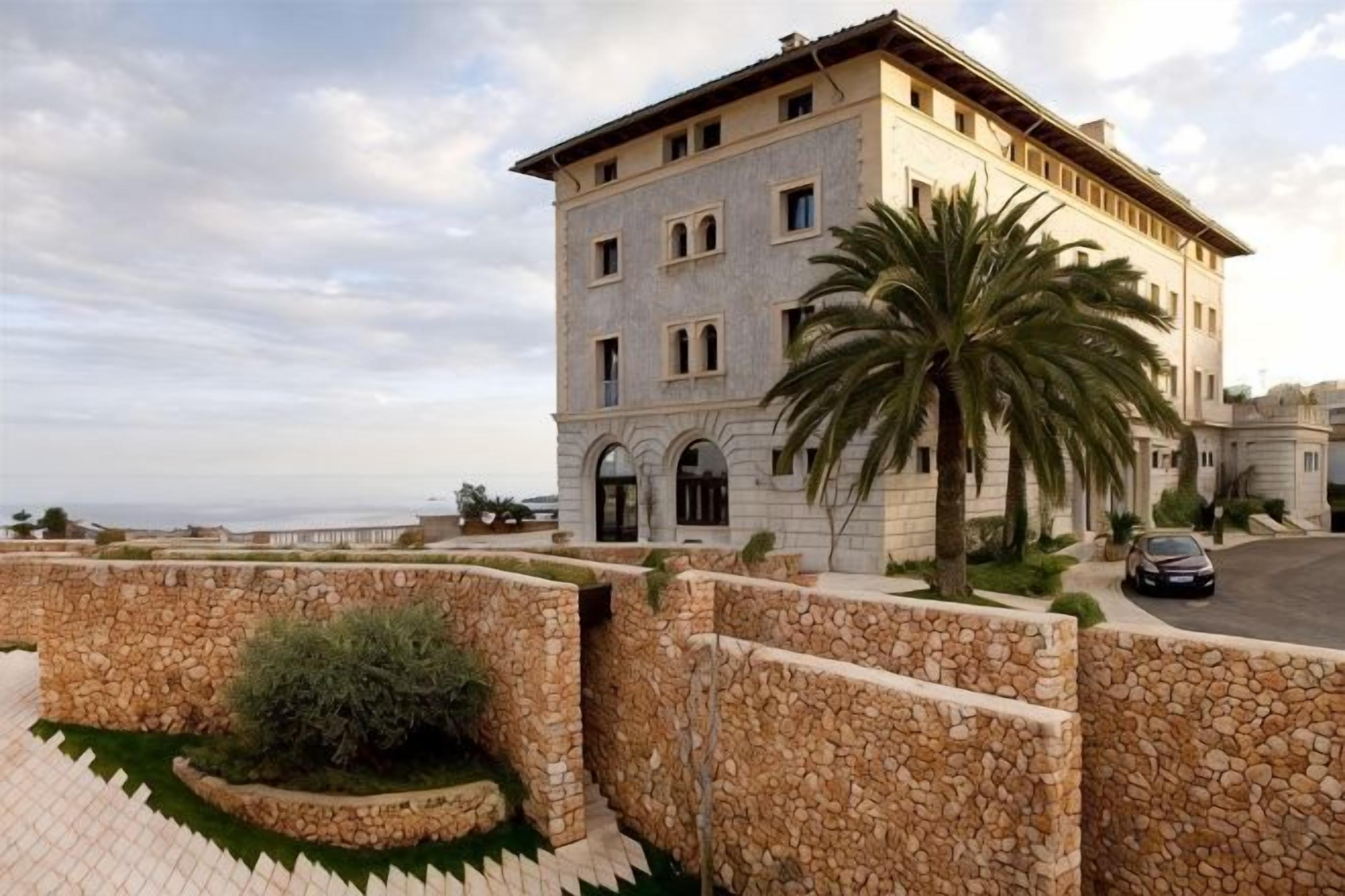 A multi-story building with a stone facade, a palm tree in the foreground, and a paved area with a stone wall.