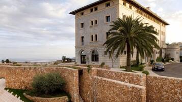 A multi-story building with a stone facade, a palm tree in the foreground, and a paved area with a stone wall.